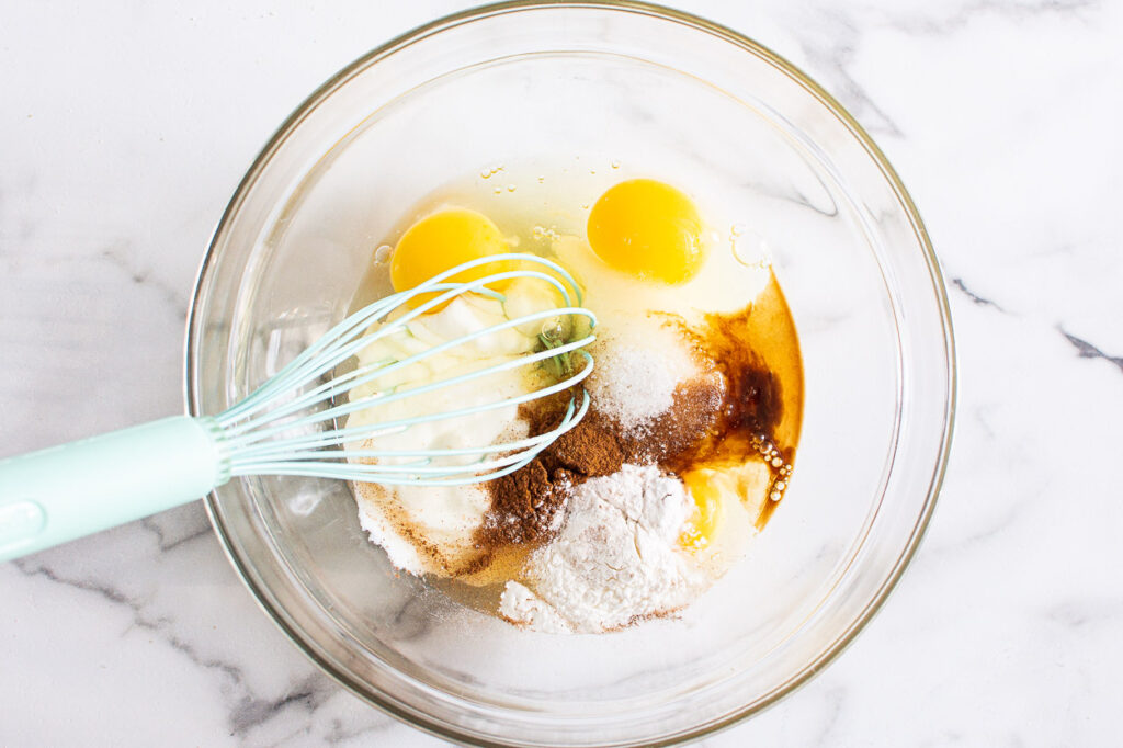 Eggs, yogurt, maple syrup and baking staples in glass bowl with a whisk. 