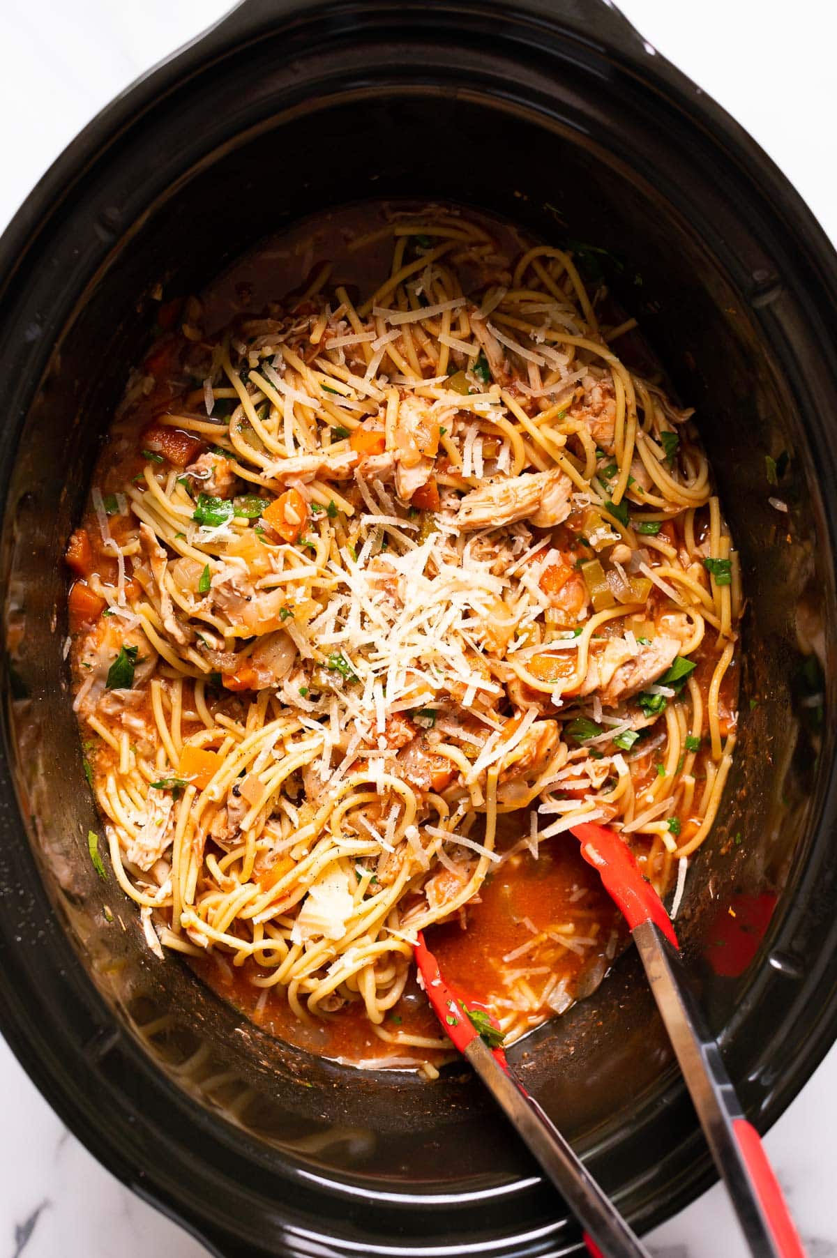 Crockpot chicken spaghetti with cheese and parsley on top and tongs inside.