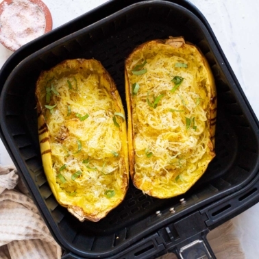 Two halves of spaghetti squash with cheese and basil in air fryer basket.