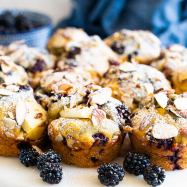 Blackberry muffins with cream cheese, sliced almonds and icing sugar served on a platter with fresh blackberries around them.