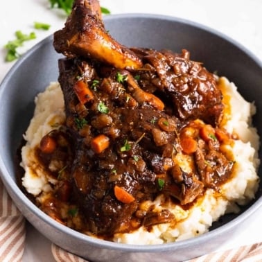 Two braised lamb shanks served over mashed potatoes with the sauce in a bowl.
