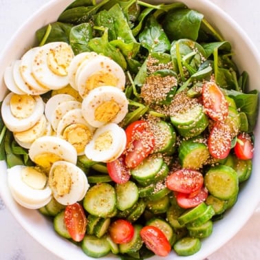A bowl of spinach salad with hard boiled eggs, tomatoes, cucumbers and sesame seeds.
