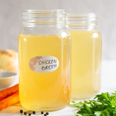 Chicken broth in glass mason jars with a label. Vegetables and peppercorns on a counter.