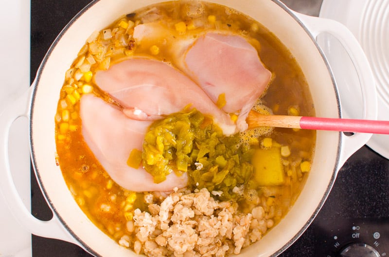 Raw chicken breasts, diced green chiles, corn, broth, mashed white beans and spices in a pot with spoon.