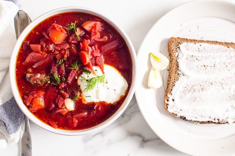 Borscht served in a bowl with yogurt, dill, garlic and rye bread on a plate.
